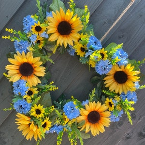 May include: A sunflower and blue flower wreath with green leaves. The wreath is hanging on a grey wooden background.