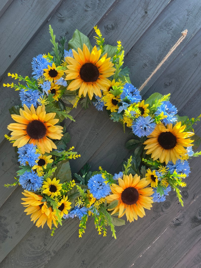 May include: A sunflower and blue flower wreath with green leaves. The wreath is hanging on a grey wooden background.