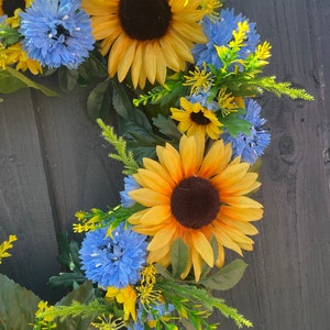 May include: A wreath made of artificial sunflowers and blue flowers. The wreath is on a dark wood background.