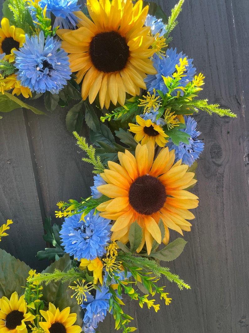 May include: A wreath made of artificial sunflowers and blue flowers. The wreath is on a dark wood background.