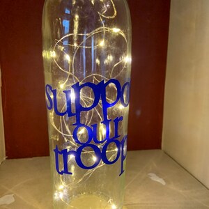 May include: A clear glass bottle with the words "Support our troops" in blue lettering. The bottle is illuminated with small, warm white LED lights. The bottle is a decorative item.