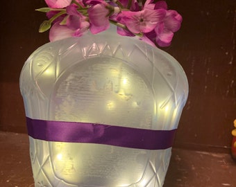 May include: A repurposed glass bottle with a purple ribbon and string lights inside. The bottle is topped with a wooden stopper and adorned with purple flowers. The bottle has a diamond pattern.