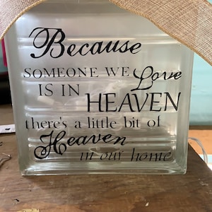 May include: A clear glass block with the words "Because someone we love is in Heaven, there's a little bit of Heaven in our home" in black script. A burlap ribbon bow adorns the top. A decorative item.