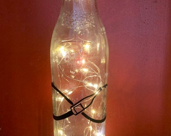 May include: A clear glass bottle with a black graphic and string lights inside. The bottle has a black design around the middle. The lights are small and warm-toned, creating a soft glow. The bottle is on a white surface.