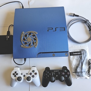 May include: A blue PS3 console with a carbon fiber design, silver fan, and the text "PS3". Includes two game controllers, a black external hard drive, a gray cable, and a package of black cables.