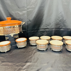 May include: A serving set featuring a large orange-brown casserole dish with a lid, set in a decorative silver-coloured metal stand. Includes eight individual ramekins with matching design. The set is designed for serving food.