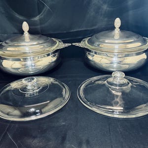Może przedstawiać: Dwie srebrne miski do serwowania z szklanymi pokrywkami. Każda miska ma srebrną pokrywkę z ozdobnym zwieńczeniem. Szklane pokrywki są owalne z małym uchwytem w kształcie gałki. Miski znajdują się na ciemnej powierzchni.