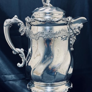Op de afbeelding: Een zilverkleurige antieke koffiepot met sierlijke details. De pot heeft een gebogen handvat, een tuit met een gebeeldhouwd gezicht en een decoratief deksel met een kleine figuur. De basis en de rand zijn ook versierd.
