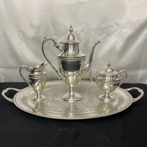 May include: A silver tea set on a silver tray. The set includes a teapot, creamer, and sugar bowl. The tray has handles and a decorative edge. The set is displayed on a black surface.
