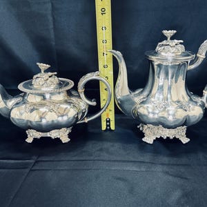 Puede incluir: Dos teteras plateadas con detalles florales y de hojas ornamentados. Las teteras tienen asas, picos y tapas decorativas. La tetera más grande mide aproximadamente 23 cm de alto, y la tetera más pequeña mide unos 15 cm de alto. Ambas se asientan sobre bases decorativas.