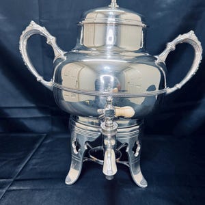 Könnte beinhalten: Eine silberfarbene Tee- oder Kaffeeserver mit verzierten Griffen und einem Zapfhahn. Der Server steht auf einem dekorativen Ständer mit drei Beinen. Der Deckel ist mit einer kleinen Verzierung versehen.