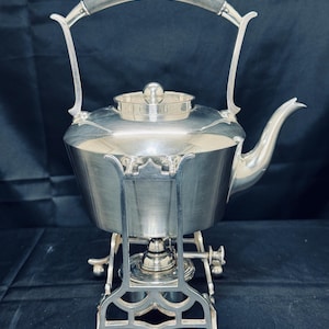 Puede incluir: Una tetera plateada con un asa oscura y un soporte a juego. La tetera tiene un pico curvo y una tapa con un pequeño pomo. El soporte tiene recortes decorativos y pequeños pies.