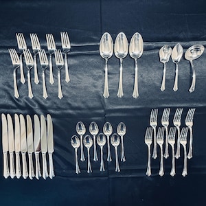 May include: An assortment of silver-colored flatware, including forks, knives, and spoons, arranged on a dark blue surface. The silverware has decorative handles and is displayed in organized rows, showcasing a complete set for dining.