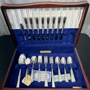 May include: A wooden box with a blue velvet interior containing a set of silver Community silverware. The silverware includes forks, knives, and spoons. The box has a label that reads "Morning Star new pattern in Community"