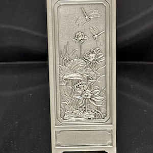 May include: A silver pewter vase with an embossed design of lotus flowers and dragonflies. The vase has a rectangular shape with a square base.