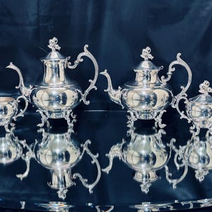 May include: A silver tea set, including a teapot, creamer, sugar bowl, and another teapot, is displayed on a reflective surface. The set has ornate handles and decorative tops. The pieces are arranged in a symmetrical pattern, reflecting their shapes.