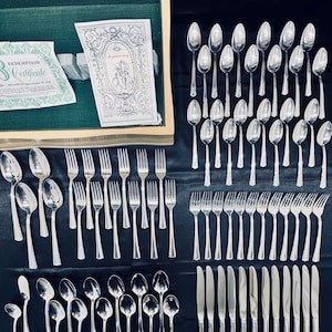 May include: A set of silverware in a silver finish, including forks, knives, and spoons. The silverware is arranged in rows on a black surface. The silverware is in a wooden box with a green lining. The box has a certificate that says "Redemption Certificate".