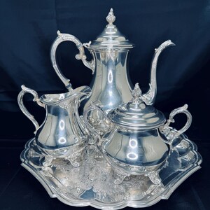 Può includere: Un servizio da tè in argento composto da una teiera, una lattiera e una zuccheriera, presentato su un vassoio decorativo. Il set presenta manici ornati e una superficie riflettente, ideale per servire tè o caffè. Il vassoio ha un motivo floreale dettagliato.