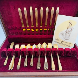 May include: A vintage silverware set in a wooden case with a red velvet interior. The set includes gold-colored knives, forks, and spoons. The case is labeled "International Sterling" and includes a card that reads "This is the era of Goldenware."