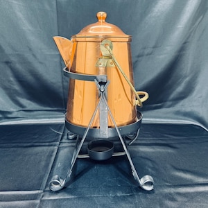 May include: A copper coffee pot with a lid and spout, on a black metal stand. The pot has a handle and a brass-coloured arm. The stand has three legs and a small circular base. A vintage kitchenware item.