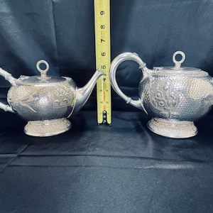 Könnte beinhalten: Zwei silberne Teekannen mit aufwendigen floralen und geometrischen Mustern. Jede Teekanne hat einen gebogenen Griff, einen Ausguss und einen Deckel mit einem kleinen Ring. Die Teekannen sind etwa 13 cm hoch und stehen auf einem dekorativen Sockel.