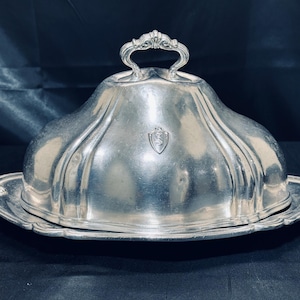 May include: Silver-plated serving dish with a domed lid and ornate handle. The dish sits on a matching oval tray with a decorative edge. A small emblem is centered on the lid. The dish is likely used for serving food.