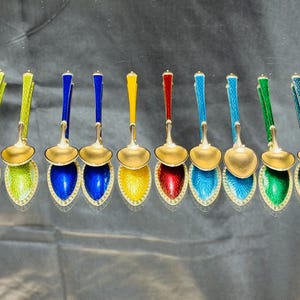 Könnte beinhalten: Eine Sammlung verzierter Löffel mit goldfarbenen Schalen und Griffen in verschiedenen Farben, darunter Grün, Blau, Gelb und Rot. Die Löffel haben dekorative, juwelenfarbene Emaille-Designs auf den Griffen und Schalen.