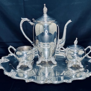 Puede incluir: Un juego de té plateado con tetera, crema y azucarero sobre una bandeja decorativa. El juego tiene detalles ornamentados y una superficie reflectante. La bandeja ovalada tiene un borde festoneado. El fondo es oscuro.