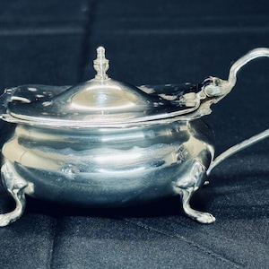 May include: A silver salt cellar with a hinged lid and three feet. The lid has a small knob on top.