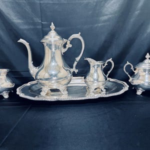 May include: A silver tea set comprising a teapot, creamer, sugar bowl, and a serving tray. The set features intricate detailing and is presented against a dark backdrop. The teapot has a tall, curved spout and a decorative lid. The creamer and sugar bowl have handles.