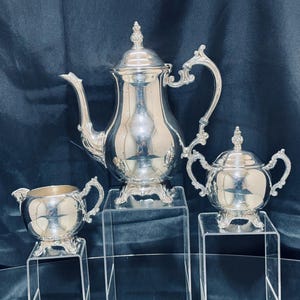 May include: A silver tea set including a teapot, sugar bowl, and creamer. The set is displayed on clear acrylic stands against a dark blue backdrop. The teapot has a curved spout and ornate handle.