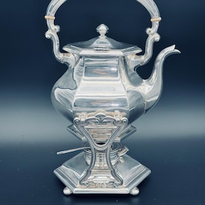 May include: A silver-colored tea kettle with a curved handle and a decorative base. The kettle has a spout and a lid with a small finial. The base has an ornate design. The kettle is likely made of metal.