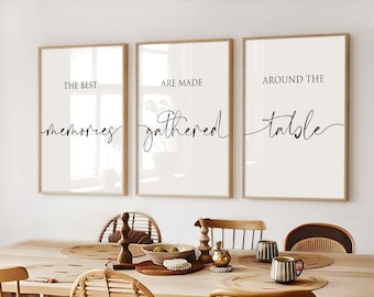 May include: Three framed prints with white backgrounds and black calligraphy text. The text reads "The best memories are made around the table". The prints are hanging on a white wall above a wooden dining table with four chairs.