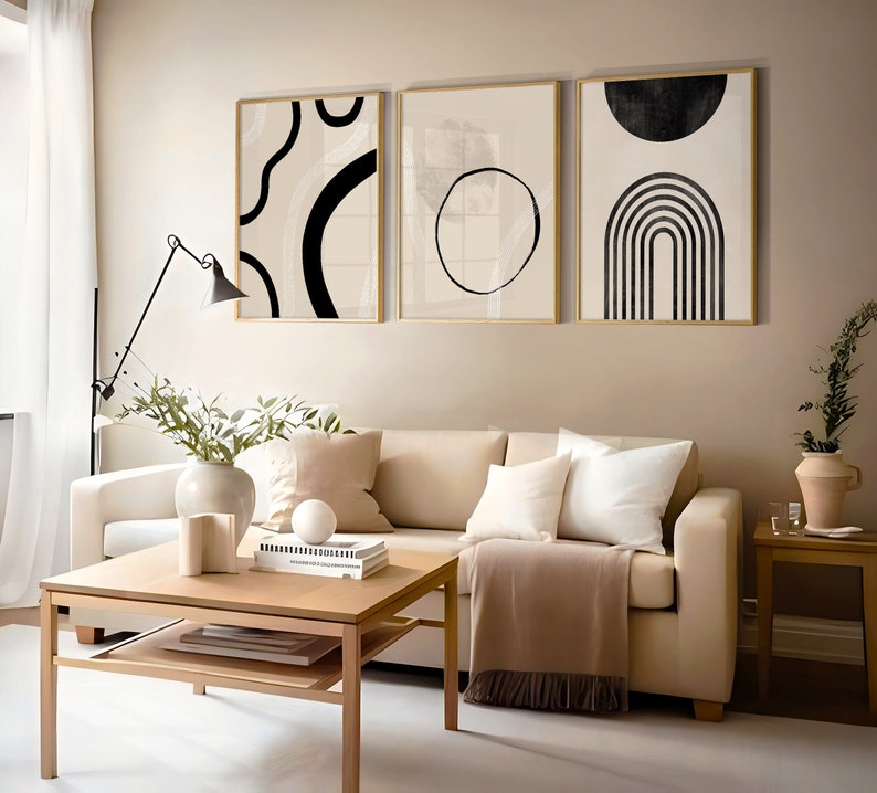 May include: Three abstract black and white art prints with geometric shapes and lines in gold frames hanging on a beige wall above a beige sofa with pillows and a coffee table.