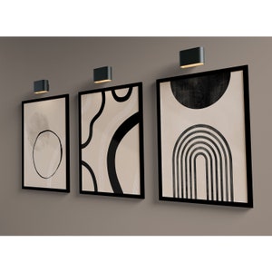 May include: Three black framed abstract art prints with beige backgrounds and black geometric shapes. The prints are hung on a beige wall with black wall sconces.