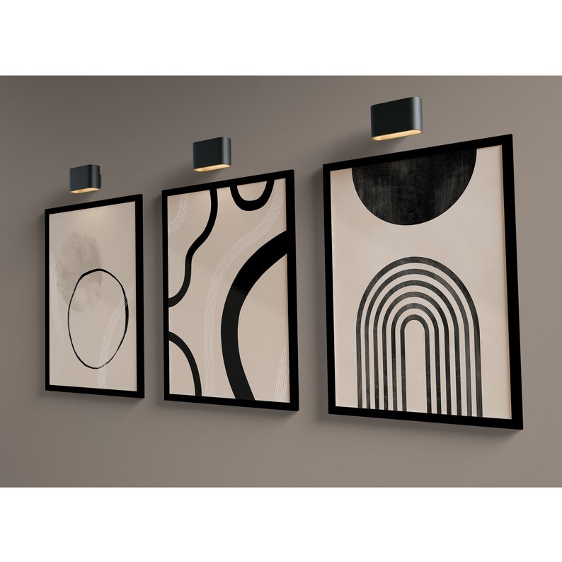 May include: Three black framed abstract art prints with beige backgrounds and black geometric shapes. The prints are hung on a beige wall with black wall sconces.