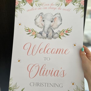 May include: A baby shower sign with a watercolor illustration of a gray elephant, surrounded by floral elements and bees. The sign reads "Welcome to Olivia's Christening 31.07.22" and "Even the smallest one can change the world."