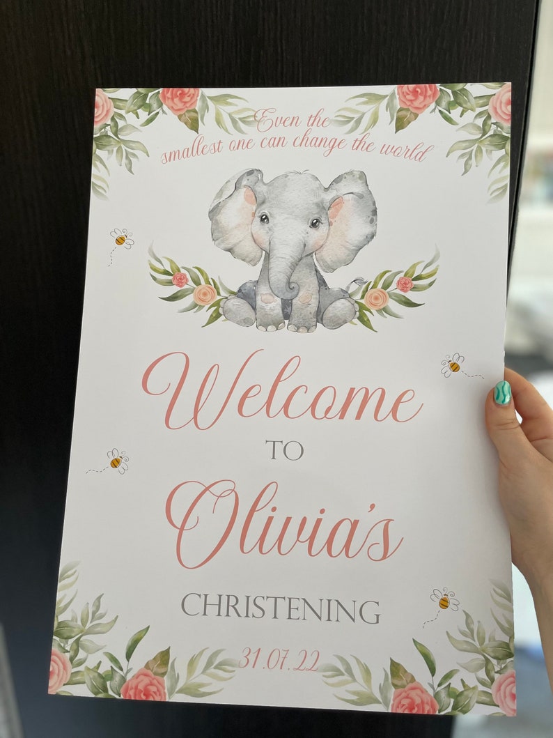 May include: A baby shower sign with a watercolor illustration of a gray elephant, surrounded by floral elements and bees. The sign reads "Welcome to Olivia's Christening 31.07.22" and "Even the smallest one can change the world."