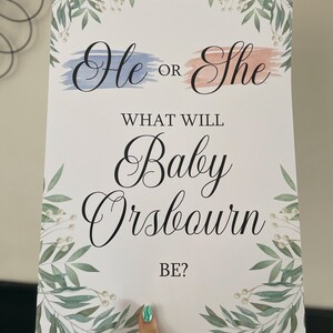 May include: A gender reveal sign with a blue and pink watercolor background. The sign says "He or She? What will Baby Orsbourn be?" with a leafy green border.