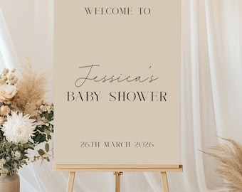 May include: A beige sign on a wooden easel with the text "WELCOME TO Jessica's BABY SHOWER 26TH MARCH 2026". The sign is surrounded by floral arrangements and a white curtain backdrop.