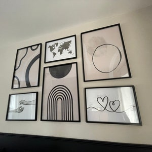 May include: A gallery wall with black framed prints featuring abstract designs, a world map, and a simple line drawing of two hearts. The prints are arranged in a symmetrical pattern on a white wall.