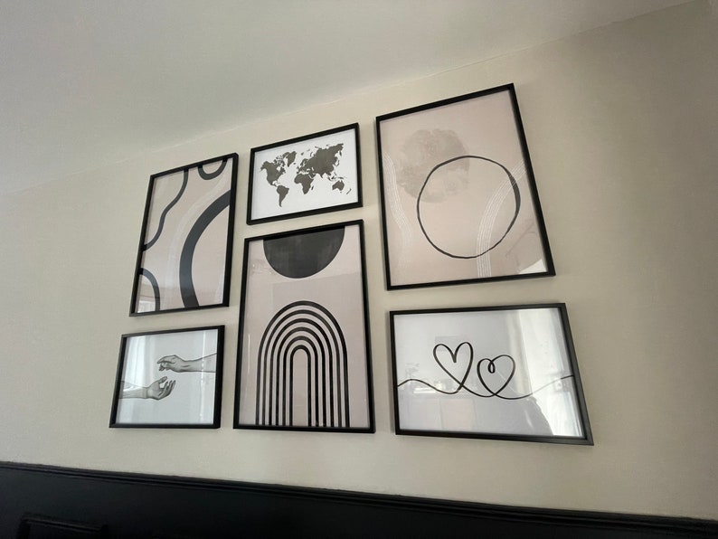 May include: A gallery wall with black framed prints featuring abstract designs, a world map, and a simple line drawing of two hearts. The prints are arranged in a symmetrical pattern on a white wall.