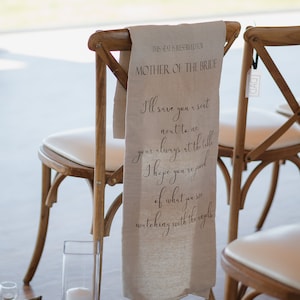 May include: A wooden chair with a white fabric sash that reads "THIS SEAT IS RESERVED FOR MOTHER OF THE BRIDE I'll save you a seat next to me your always at the table I hope you're proud of what you see watching with the angels."