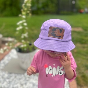 Può includere: Un cappello a secchiello lavanda con una toppa in pelle marrone con un design stilizzato. Il cappello è indossato da una persona che indossa una maglietta rosa con la parola "niks" in un carattere giocoso. Lo sfondo è una scena all'aperto sfocata.