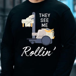 May include: Black long-sleeve t-shirt with a white graphic of a cartoon robot with the text "They See Me Rollin'"