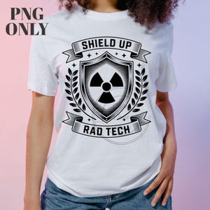 Peut inclure: T-shirt blanc avec un motif noir et blanc. Le motif comprend un bouclier avec un symbole de radiation, le texte "SHIELD UP" au-dessus et "RAD TECH" en dessous. Des feuilles de laurier encadrent le bouclier.