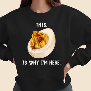 Könnte beinhalten: Schwarzes Sweatshirt mit dem Text "THIS. IS WHY I'M HERE." In der Mitte befindet sich eine Grafik eines gefüllten Eies. Das Ei hat eine weiße Basis mit gelber Füllung und einer Prise Paprika.