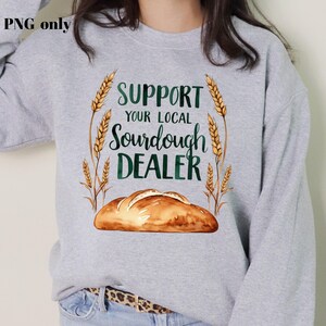 Op de afbeelding: Een grijs sweatshirt met de tekst "Support Your Local Sourdough Dealer" in groen en bruin. De tekst wordt omringd door tarwehalmen en een zuurdesembrood.