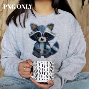 Könnte beinhalten: Ein hellgraues Sweatshirt mit einem Cartoon-Waschbären in Jeanskleidung. Der Waschbär hat große, ausdrucksstarke Augen und ein blau-graues Farbschema. Die Person hält einen weißen Becher mit einem schwarzen Punktmuster. Der Text "PNG ONLY" befindet sich oben links.