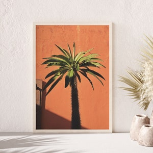 May include: A framed print of a green palm tree against a bright orange wall. The print is in a white frame and is sitting on a white surface. There are two beige vases with dried plants in front of the print.
